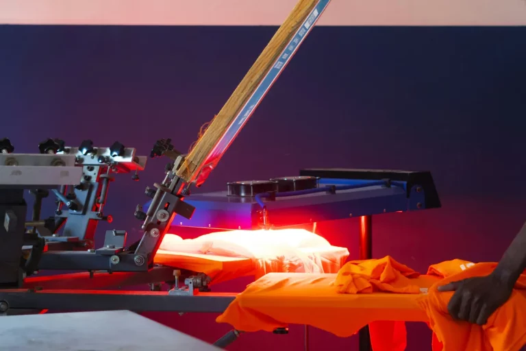 Atelier de sérigraphie à Dakar : un ouvrier positionne des t-shirts orange sous une machine d’impression illustrant le nouveau code du travail décrypté par Novallia